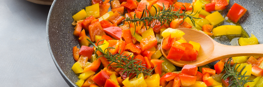 Close up of red, yellow, and orange bell peppers in a pan with a sprig of rosemary.