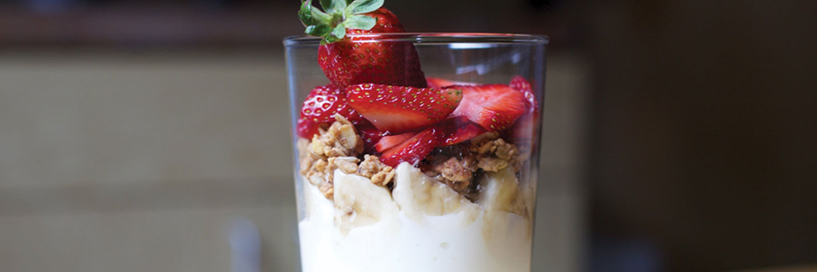 View of clear stemmed glass filled with layers of yogurt, bananas, granola, and strawberries.