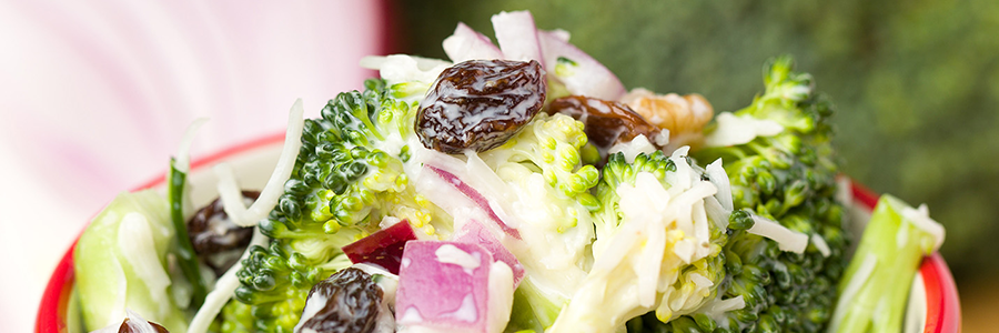 Close up view of a salad with broccoli florets, chopped red onion, shredded cheese, and raisins in a colorful red and white striped bowl.