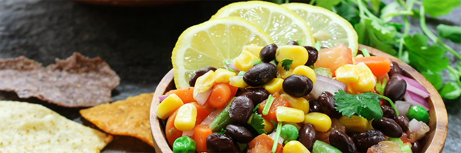 Close up shot of salsa with black beans, corn, and tomatoes in a wood bowl. Salsa is garnished with lemon slices.