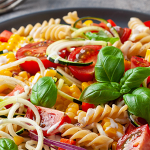 Close up of a plate with rotini pasta, corn, cherry tomatoes, and zucchini spirals. Plate is garnished with basil leaves.