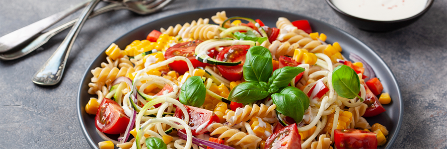 Close up of a plate with rotini pasta, corn, cherry tomatoes, and zucchini spirals. Plate is garnished with basil leaves.