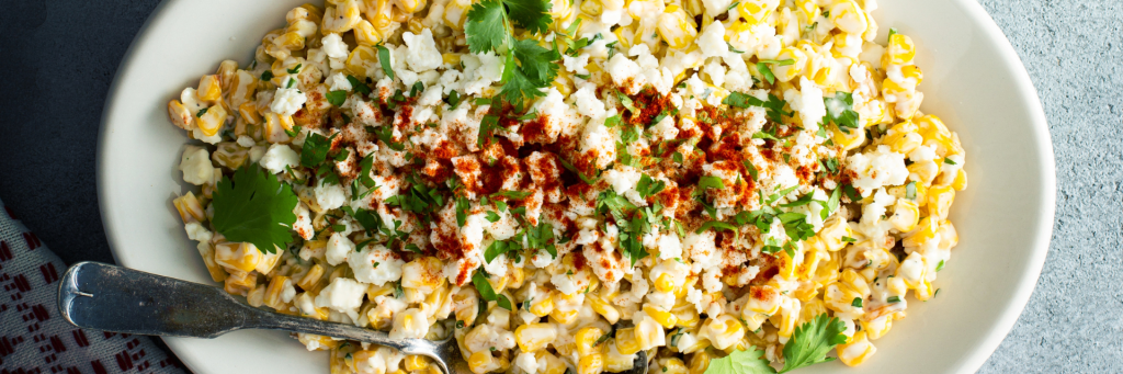 Close up of a white platter with creamy corn salad with crumbled Cotija cheese and creamy dressing. Garnished with Chipotle seasoning and cilantro.
