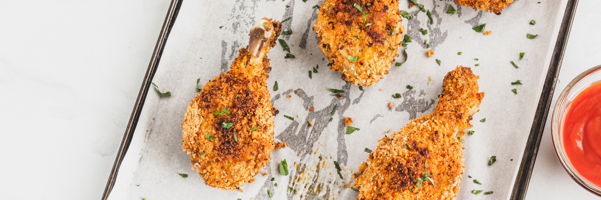 Wide shot of breaded chicken drumsticks on a parchment -lined baking sheet. Chicken is garnished with parsley.