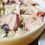 Two bowls set on a table; one larger bowl with apples, celery, and raisins coated in yogurt. The second bowl is much smaller and filled with chopped pecans.