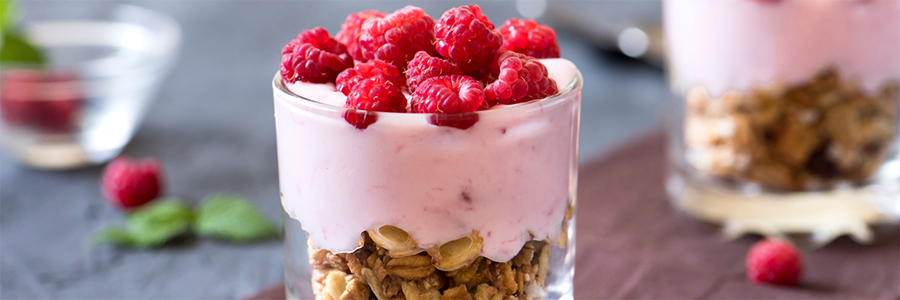 Wide shot of two glasses layered with granola, strawberry yogurt and topped with fresh raspberries.