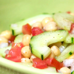 Close up view of a salad with sliced cucumbers, diced tomatoes, onions and chickpeas in a large green salad platter. Salad is garnished with black pepper.