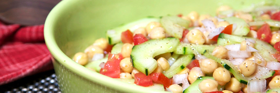 Close up view of a salad with sliced cucumbers, diced tomatoes, onions and chickpeas in a large green salad platter. Salad is garnished with black pepper.