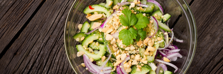 Close up of chopped cucumber, peanuts, red onion slices in a clear bowl with cilantro garnish on top.