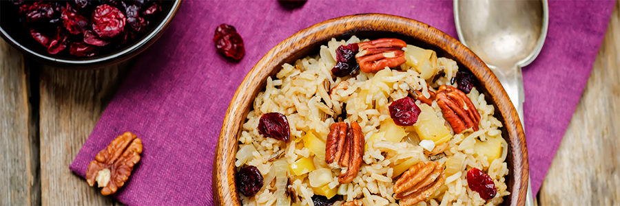 Close up of a bowl of rice with dried cranberries and pecans. Bowl is setting on a deep pink placemat and is displayed with a small bowl of dried cranberries in the upper left corner.