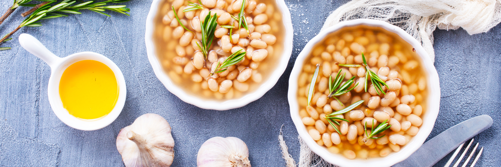 On top of a blue background, with bulbs of garlic and utensils around there is an aerial view of two white bowls filled with Great Northern beans with sprigs of greenery on top.