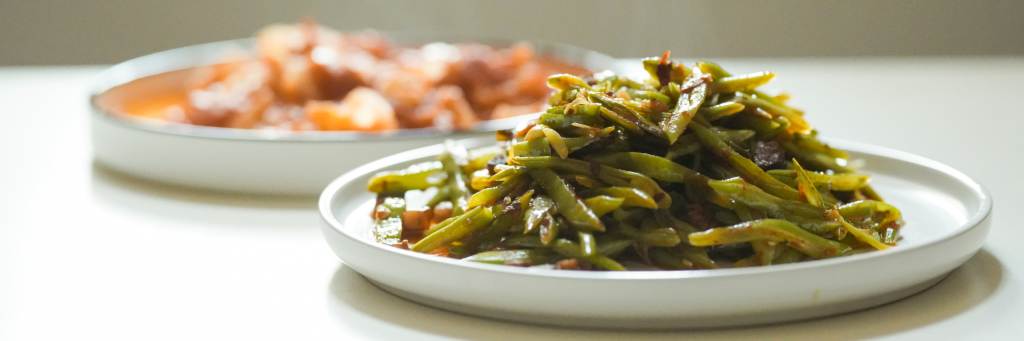 Wide shot of a plate of seasoned green beans with plate of yams blurred in the background.