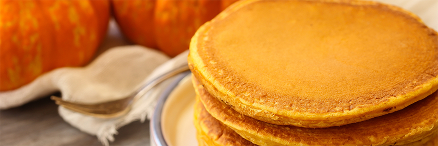 Close up shot of a stack of four pumpkin pancakes with two small pumpkins blurred in the background.