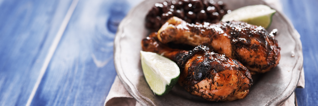Shot of two baked chicken drumsticks marinated in jerk seasoning with a side of black beans in the background. Plate is garnished with lime wedges.