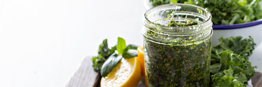 Wide shot of a small jar of pesto displayed with kale leaves and lemon. A spoon with a spoonful of pesto in the foreground.