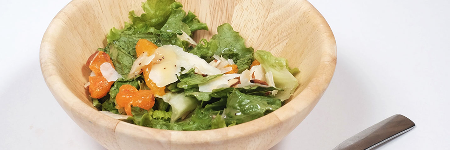 Close up view of a wood salad bowl containing a leafy green salad with mandarin oranges and almonds. Topped with poppy seed dressing and shaved Parmesan cheese.