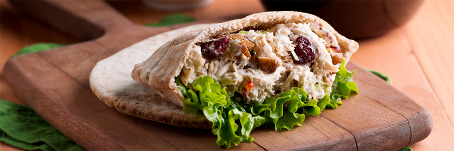 A Pear Waldorf and Chicken Stuffed Pita, with chicken, raisins, lettuce, and nuts, sitting on a medium to dark wood cutting board which is on top of a lighter wood table.