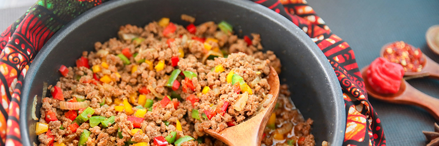 Shot of a skillet filled with a mixture of ground turkey, peppers, onions and tomatoes. Skillet is resting on a colorful African-inspired cloth. There is a wooden spoon placed inside the skillet.