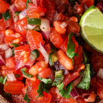 Close up of fresh salsa with tomatoes, red onion, jalapeno, and cilantro on a cutting board. Garnished with a lime wedge.