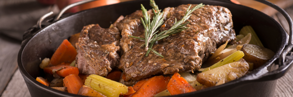 A close up of a pot roast with celery, carrots, carrots, and a sprig of rosemary on top in a black pot.