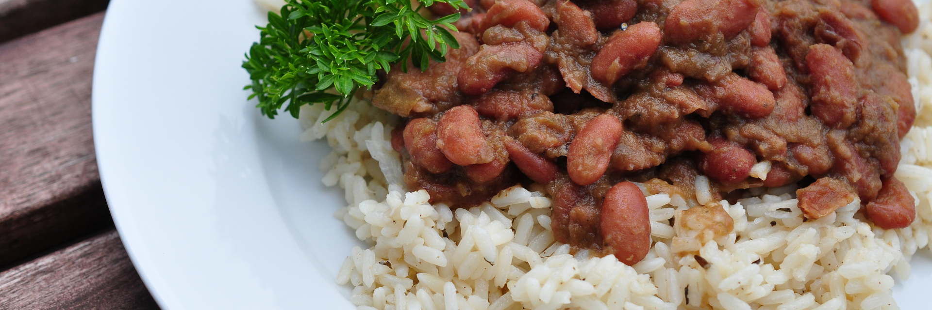 Close up of red bens, white rice, and a sprig of parsley as garnish on a white plate, on top of a wooden table.