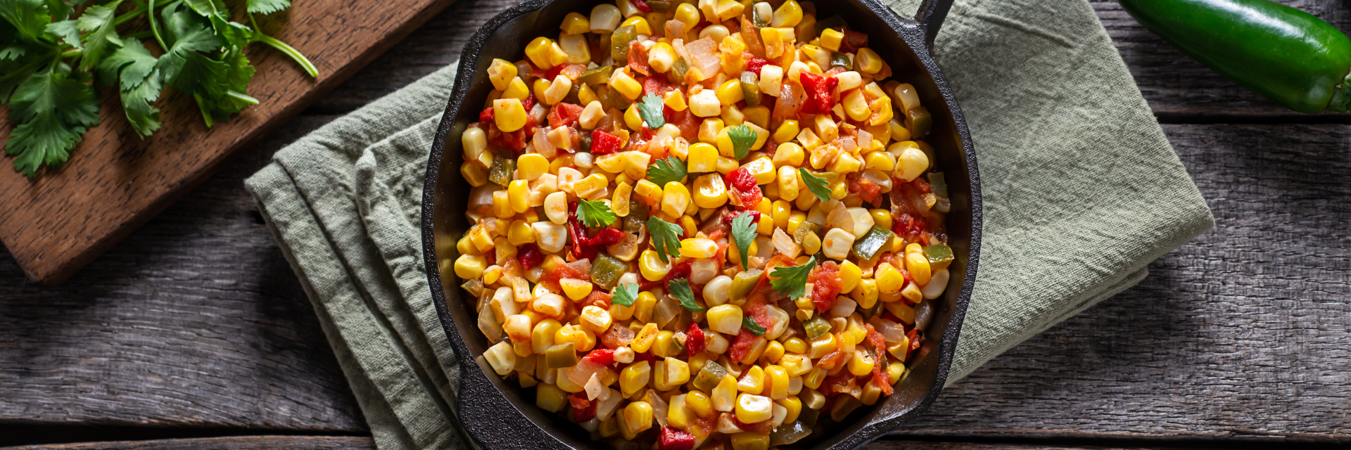 Seasoned corn in a skillet with onion, green pepper, and spices with a green towel under it sitting on a wooden table. On the left is some garnish on and cutting board and a green chili pepper on the right.