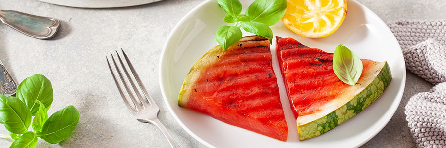 Wide shot of a plate with two slices of grilled watermelon. Plate is garnished with basil leaves and a half a lemon.