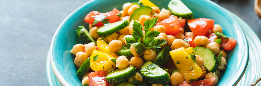 Close up shot of a colorful salad with chickpeas, yellow peppers, cucumbers and tomatoes in a light dressing. Salad is served in a blue ceramic bowl.