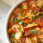 Tomato and Cabbage Soup with carrots, tomatoes, cabbage, and greenery in a white bowl with a cutting board of greenery on the left side of the photo.