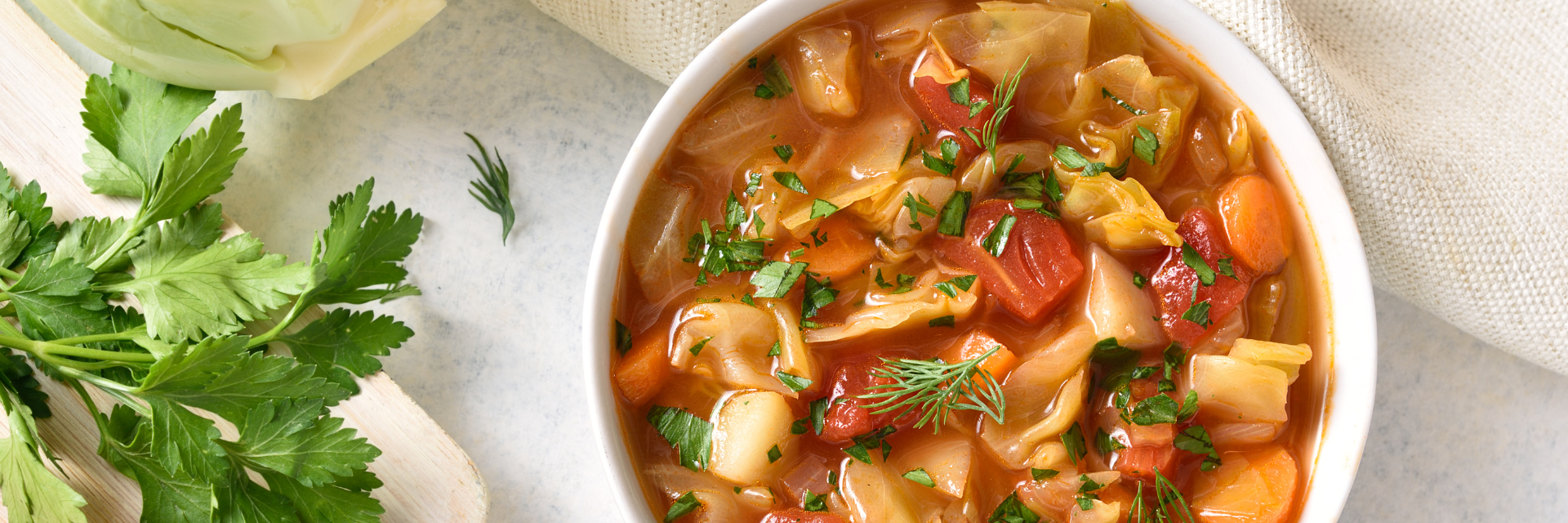 Sopa de tomate y col con zanahorias, tomates, col y verduras en un cuenco blanco con una tabla de cortar con verduras en el lado izquierdo de la foto.