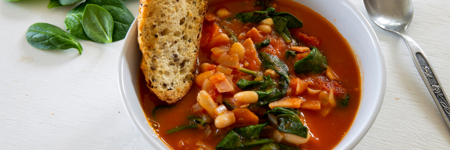 Close up of tomato based soup with white beans, tomatoes, and spinach in a white bowl. Slice of toasted bread on the side of the bowl with spinach leafs and silver spoon.