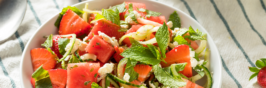 Close up of a salad with cubed watermelon, sliced strawberries, crumbled feta, and cucumber. Garnished with mint leaves.