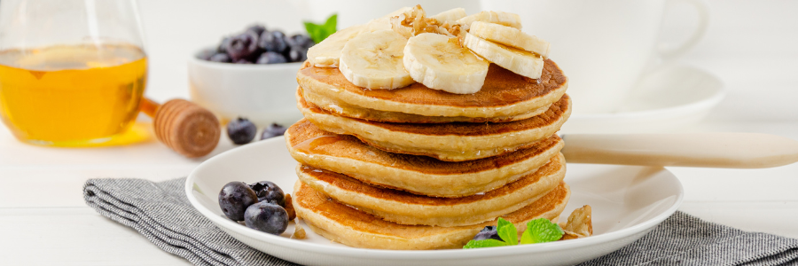 Close up of a stack of 5 pancakes with light syrup and bananas on top on a white plate with blueberries and mint to the side.