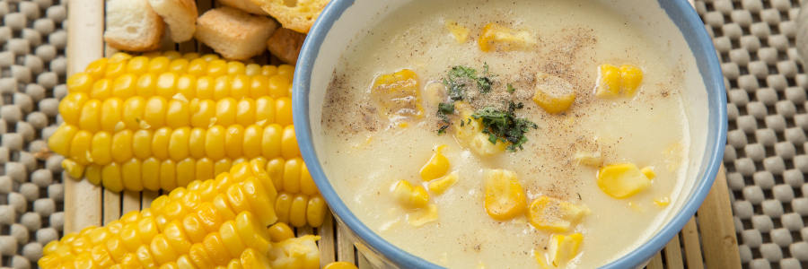 Close up of corn soup, creamy yellow with corn kernals on top in a bowl. Cooked corn and bread to the side.