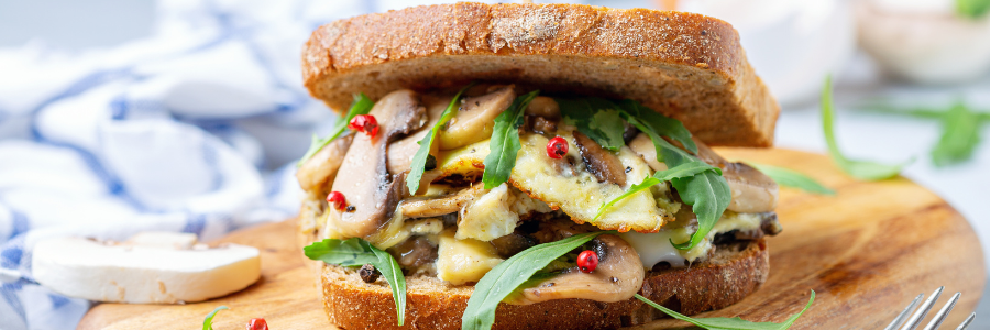 Close up of a sandwich made of eggs, mushrooms, and arugula on whole wheat bread, resting on a wooden cutting board. Fork and butter knife to the side.