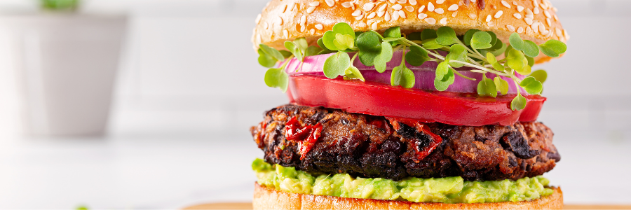 Close up of a burger with a black bean and beef patty, tomato, guacamole, sprouts, and red onion on a bun.