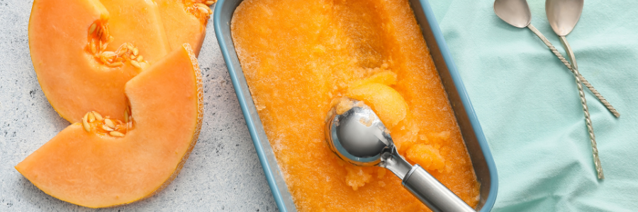Closeup of a small pan of frozen melon sorbet, orange in color with ice cream scoop. Orange melon to the side.