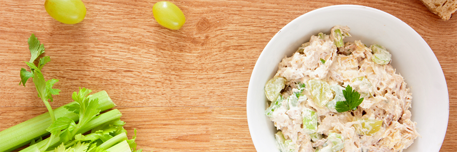 Wide shot of a bowl of chicken salad with green grapes, onion, and celery in a creamy dressing. Bowl is on a wooden table with two grapes and a celery bunch.