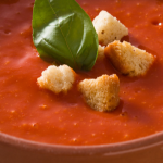 Close up of tomato soup in beige bowl with croutons and basil on top.