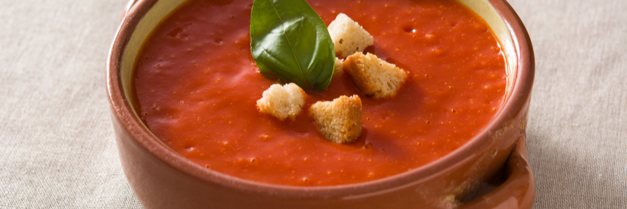Close up of tomato soup in beige bowl with croutons and basil on top.