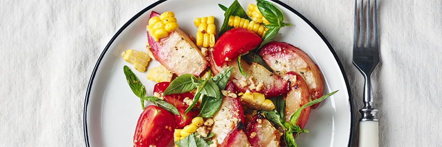 Shot of a salad with sliced peaches, fresh cut corn, quartered cherry tomatoes and basil leaves on a white plate with black trim.