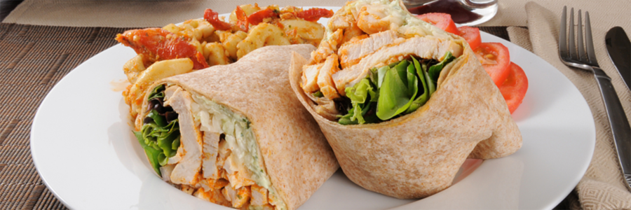 Wide shot of a sandwich wrap with chicken and veggies wrapped in a whole wheat tortilla. Sandwich wrap is cut into two and displayed on a white plate with tomato slices on the side as garnish.