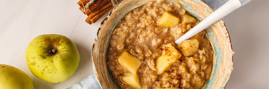 Wide shot of a bowl of oatmeal with diced apples and cinnamon. Bowl is displayed with a whole golden delicious apple and decorative cinnamon sticks on the table.