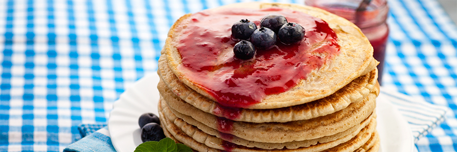 Shot of a stack of pancakes drizzled with raspberry sauce and topped with whole blueberries. Plate is garnished with mint leaves and additional blueberries.
