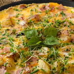 Wide shot of a frittata with ham, potatoes, and herbs in a cast iron skillet. Frittata is garnished with a variety of herbs and is displayed on a table with chives as decor.