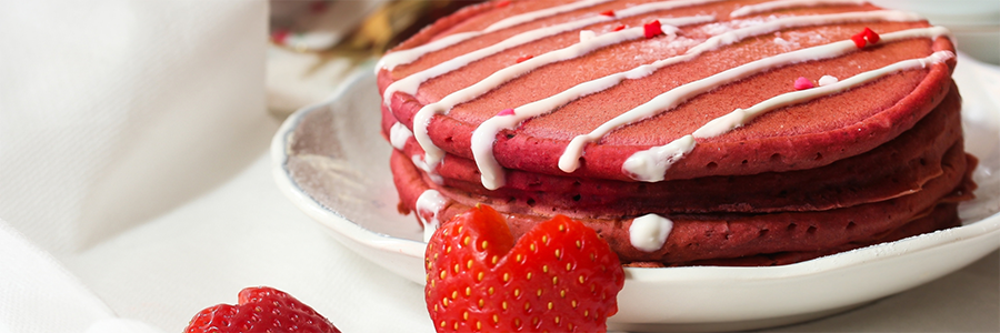 Wide shot of a small stack of pink pancakes drizzled with Greek yogurt and garnished with heart-shaped strawberry slices.