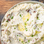 Wide shot of a bowl of mashed red potatoes topped with melted butter and chopped fresh parsley.