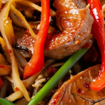 Close up shot of a stir fry with strips of pork tenderloin, red bell pepper, and green onion with noodles in a chili-garlic sauce.