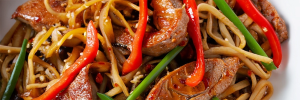 szechuan-pork Close up shot of a stir fry with strips of pork tenderloin, red bell pepper, and green onion with noodles in a chili-garlic sauce.