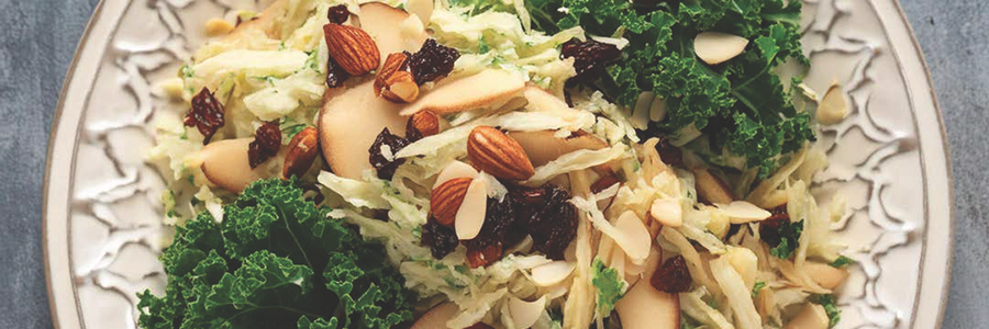 Close up shot of a slaw with shredded rutabaga, apples, pears, and kale. Slaw is garnished with dates and almonds.